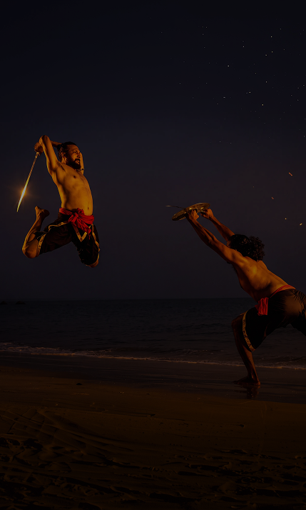 Two shirtless men performing martial arts on a beach at night, one jumping mid-air holding a sword and the other crouched holding a shield.