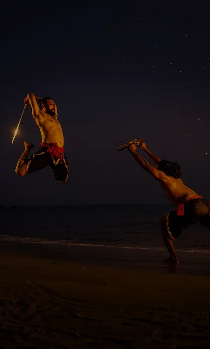 Two shirtless men performing martial arts on a beach at night, one jumping mid-air holding a sword and the other crouched holding a shield.
