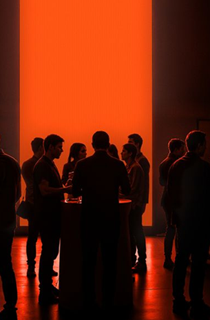 Silhouettes of people standing and talking in a dimly lit room with a large bright orange rectangular light in the background.
