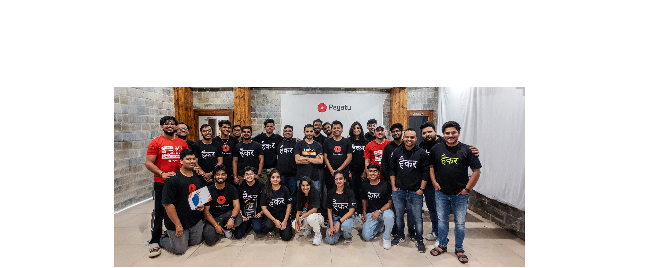 Group photo of 26 people standing and kneeling indoors in front of a Payatu banner, most wearing black shirts or hoodies with Hindi text, two in red shirts, some holding certificates and a trophy.