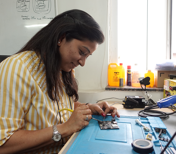 A woman wearing a yellow and white striped shirt working intently on an electronic circuit board at a workbench with tools and equipment, including tape, wires, and a soldering kit, under natural light from a window.