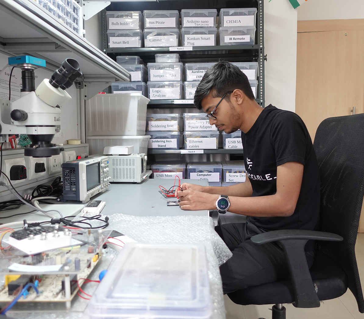 A man wearing glasses and a black t-shirt is seated at a workstation in a electronics lab, working with wires connected to a small circuit board. Behind him are shelves with labeled clear plastic bins containing electronic components. On the desk are electronic test instruments including an oscilloscope and a microscope.