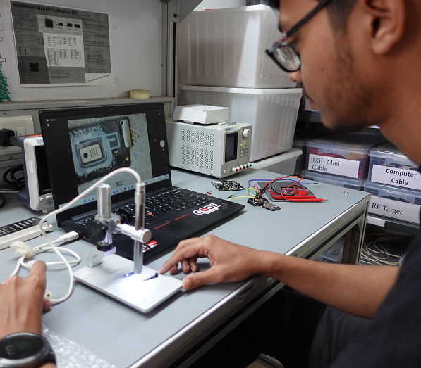 Person working at a desk with a laptop showing a magnified close-up of an electronic chip, using a microscope setup with connected cables and testing equipment nearby labeled USB Mini Cable, Computer Cable, and RF Target.