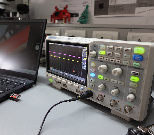 Digital oscilloscope on a desk displaying waveform data on its screen, connected by cable to nearby laptop, with various control buttons and knobs visible on the device panel.