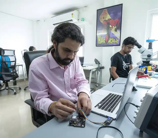 Two men working in a bright office with computers and electronic equipment; the man in the foreground is connecting wires to a circuit board, while the other man is seated at a desk with a microscope and laptop.