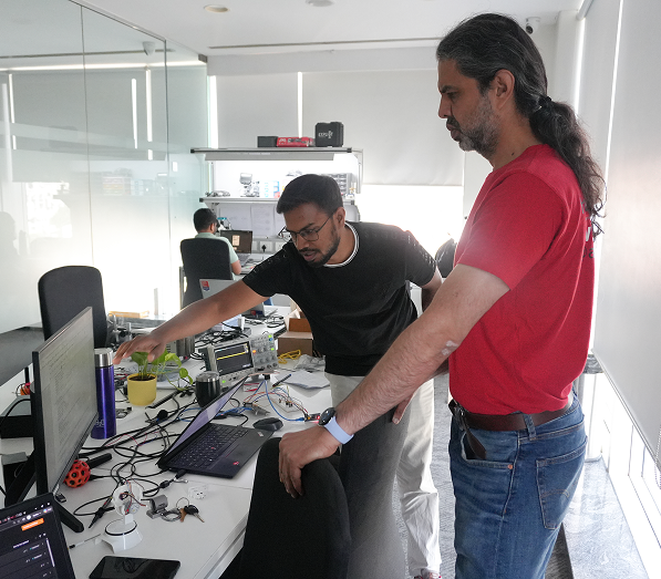 Two men working together at a cluttered desk with laptops, electronic equipment, cables, and a small plant in a bright office space with glass walls.