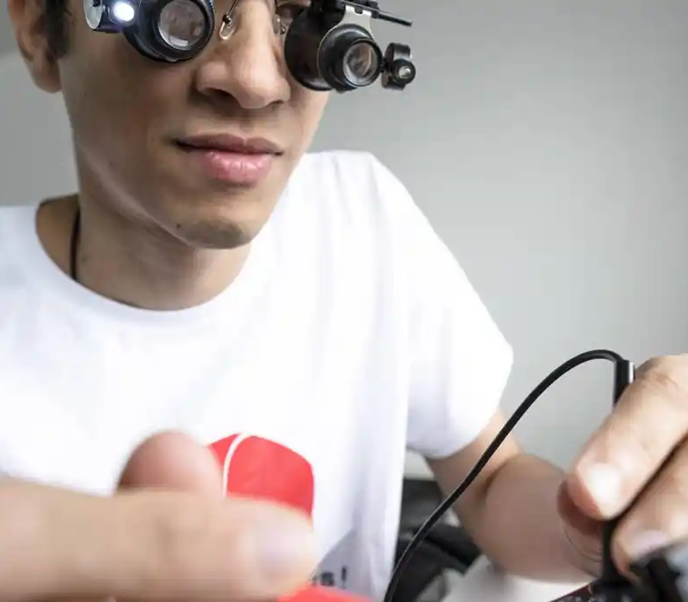 Man wearing magnifying glasses with a small light, closely examining a black cable or wire held in his hands.