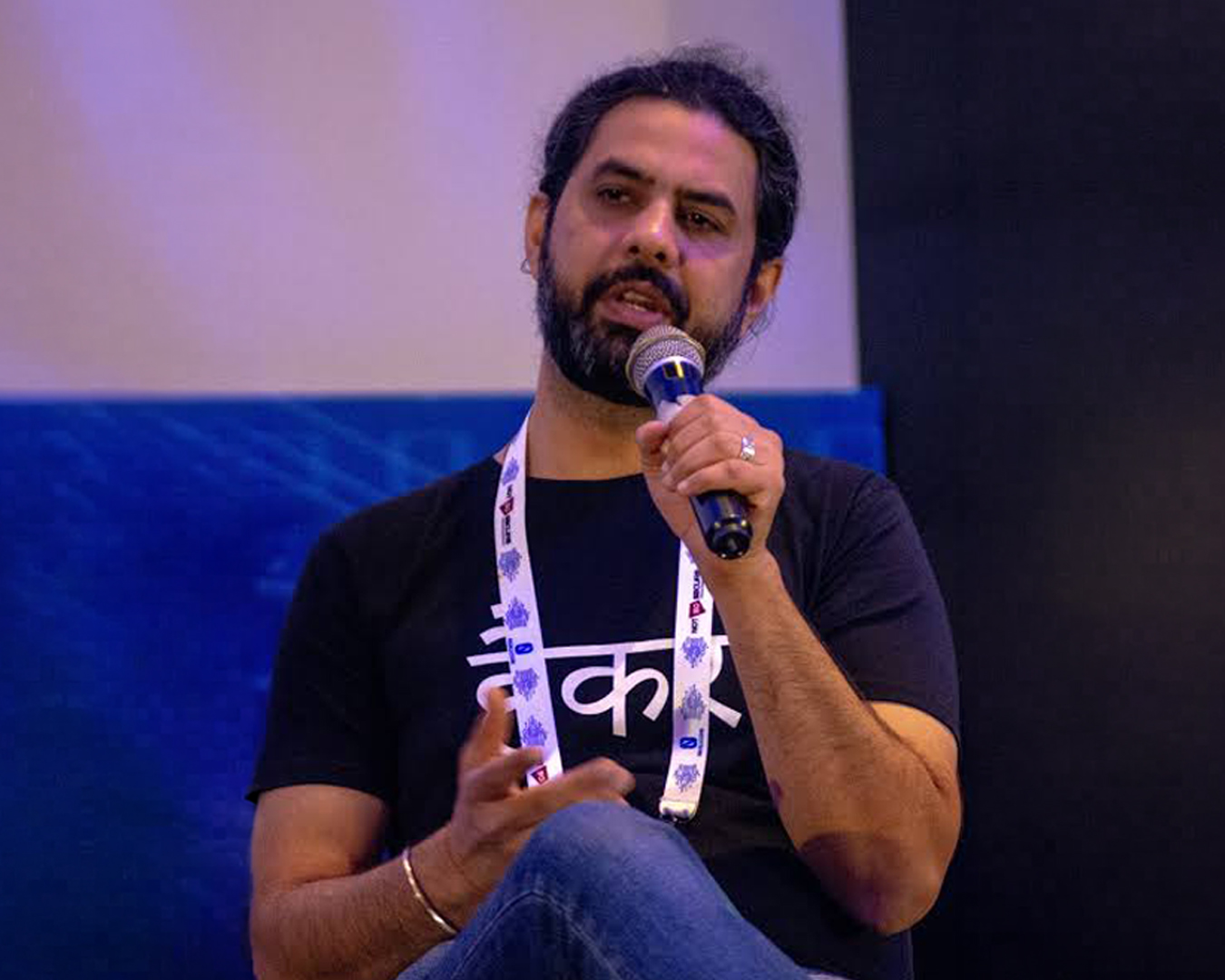 Bearded man with dark hair tied back speaking into a microphone while seated, wearing a black t-shirt with white text and a white lanyard.