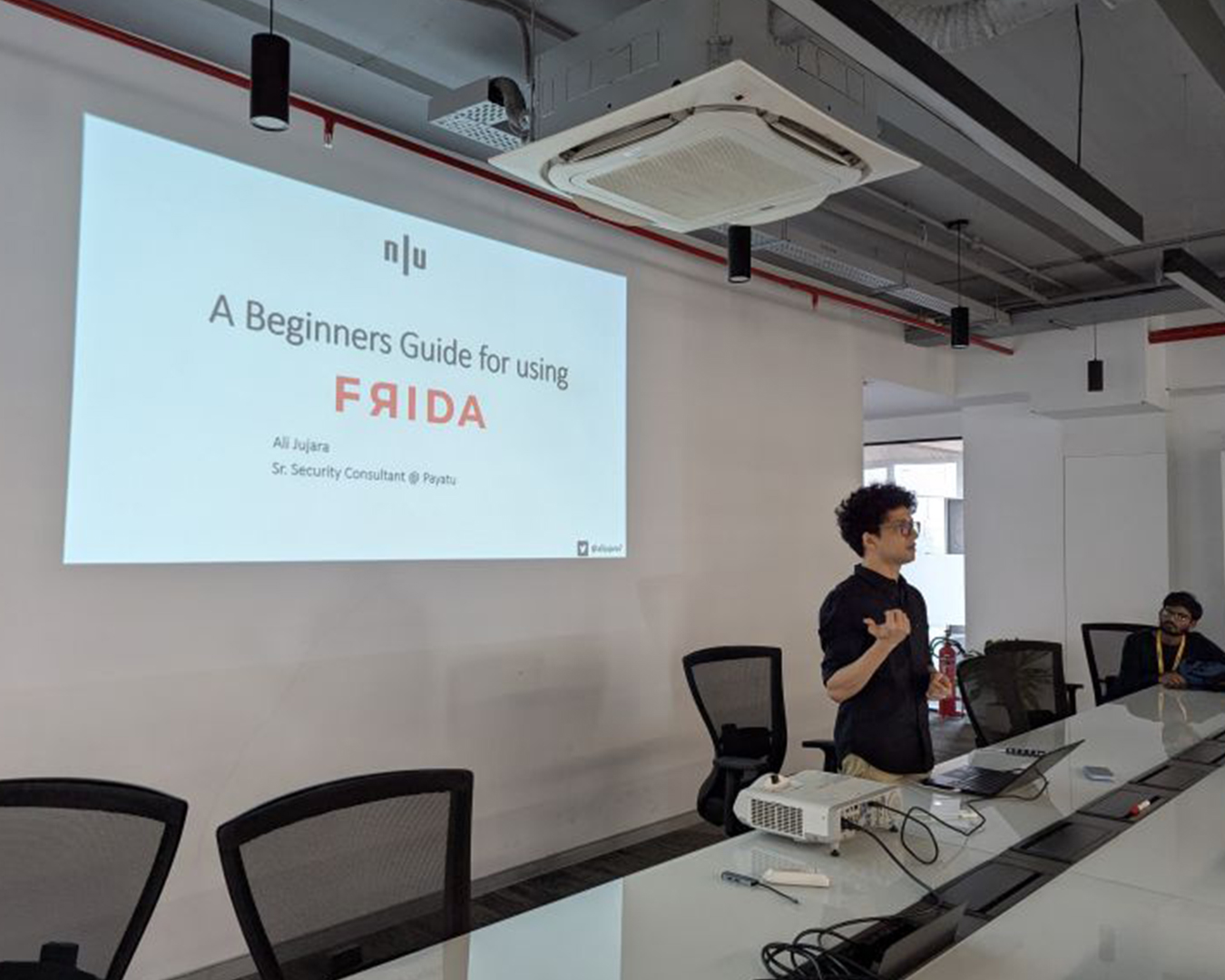 A person giving a presentation in a conference room with a slide that reads 'A Beginners Guide for using FRIDA' by Ali Jujara, Sr. Security Consultant at Payatu. The presenter stands near a white table with a projector and laptop, speaking to an audience member seated on the right.