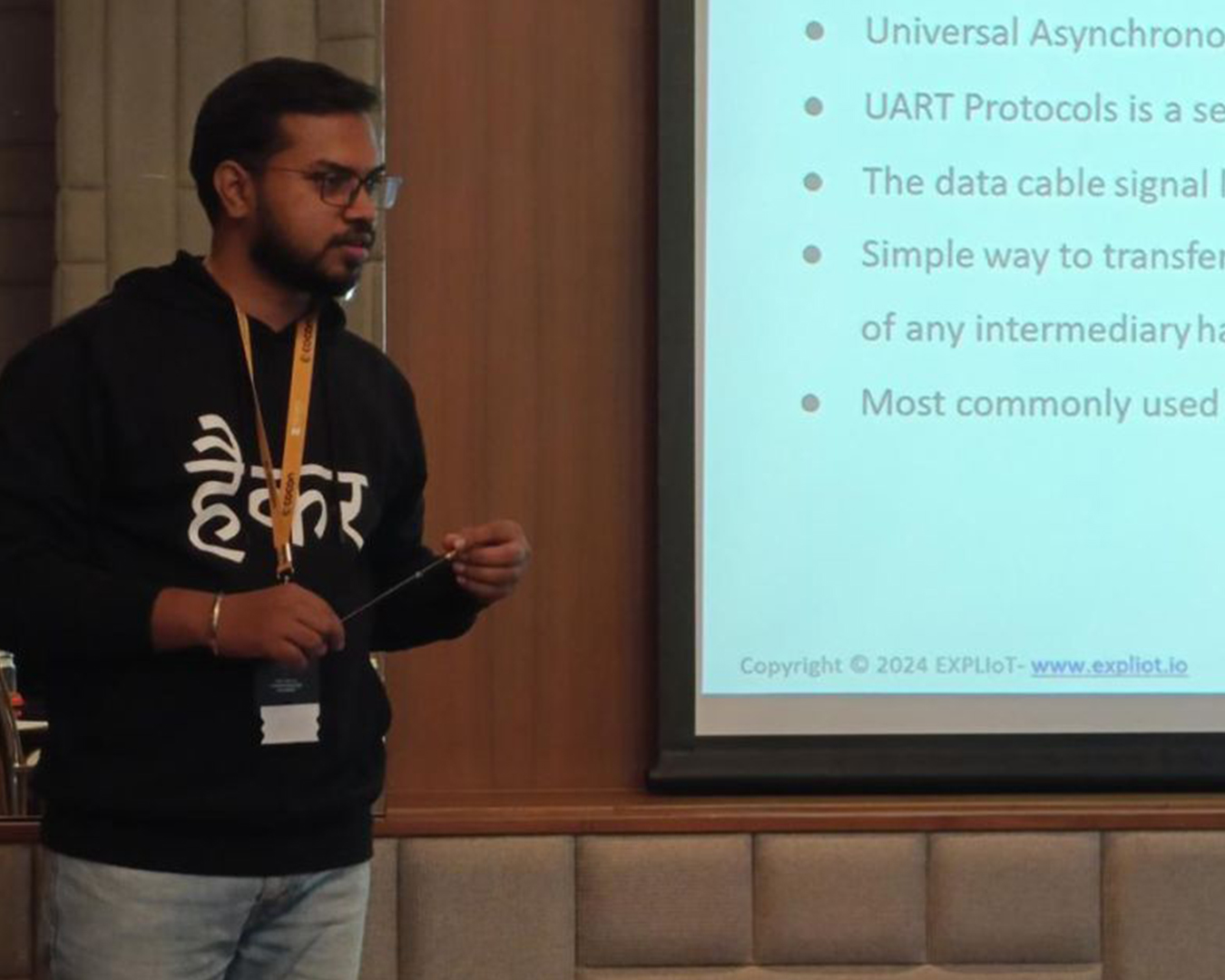 Man wearing glasses and a black hoodie with Hindi text is giving a technical presentation, standing beside a projector screen displaying bullet points about UART protocols.