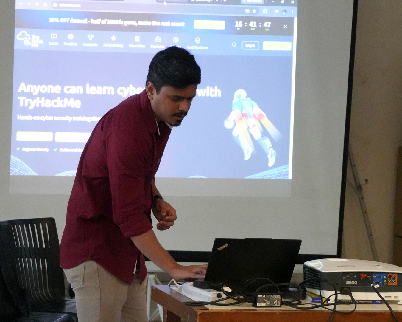 Man in a maroon shirt operating a Lenovo ThinkPad laptop on a table with a BenQ projector projecting a TryHackMe cybersecurity training website on a screen behind him.