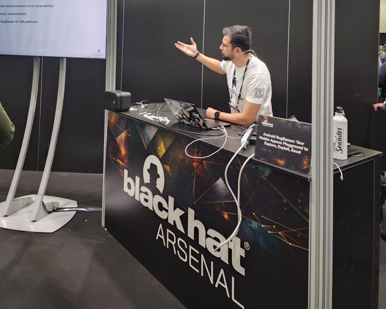 A man wearing a white t-shirt speaks and gestures while sitting behind a Black Hat Arsenal booth with a laptop, cables, and a small sign that reads 'Android BugBazaar: Your Mobile Appsec Playground to Explore, Exploit, Excel'.
