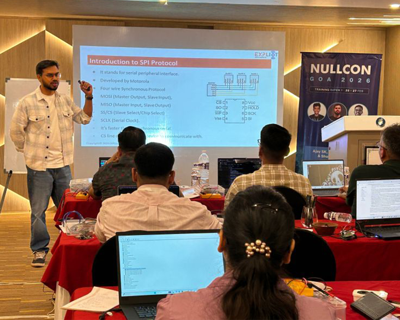 A man in a plaid shirt presents a technical lecture on SPI Protocol to a seated audience with laptops in a classroom. A popular banner reads 'NULLCON GOA 2026' alongside photos of three speakers. The projector screen displays a slide titled 'Introduction to SPI Protocol' with text and a circuit diagram.