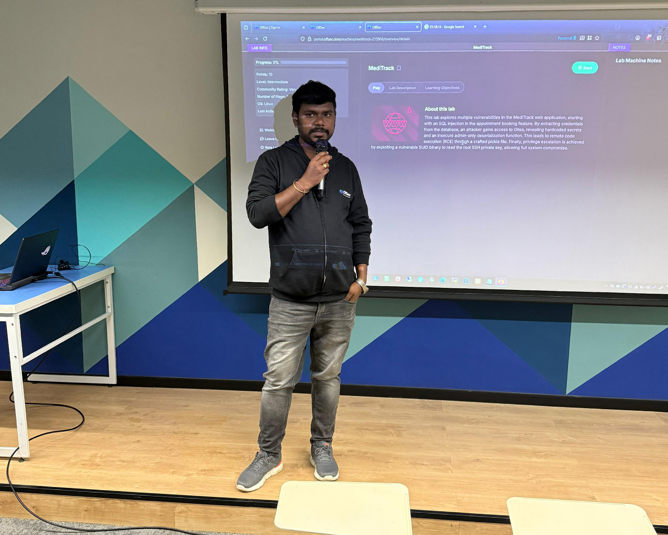 A man in a black OffSec hoodie and gray jeans holds a microphone and stands on a wooden floor in front of a projector screen displaying a Meditrack web application lab overview with details about SQL injection and privilege escalation vulnerabilities.