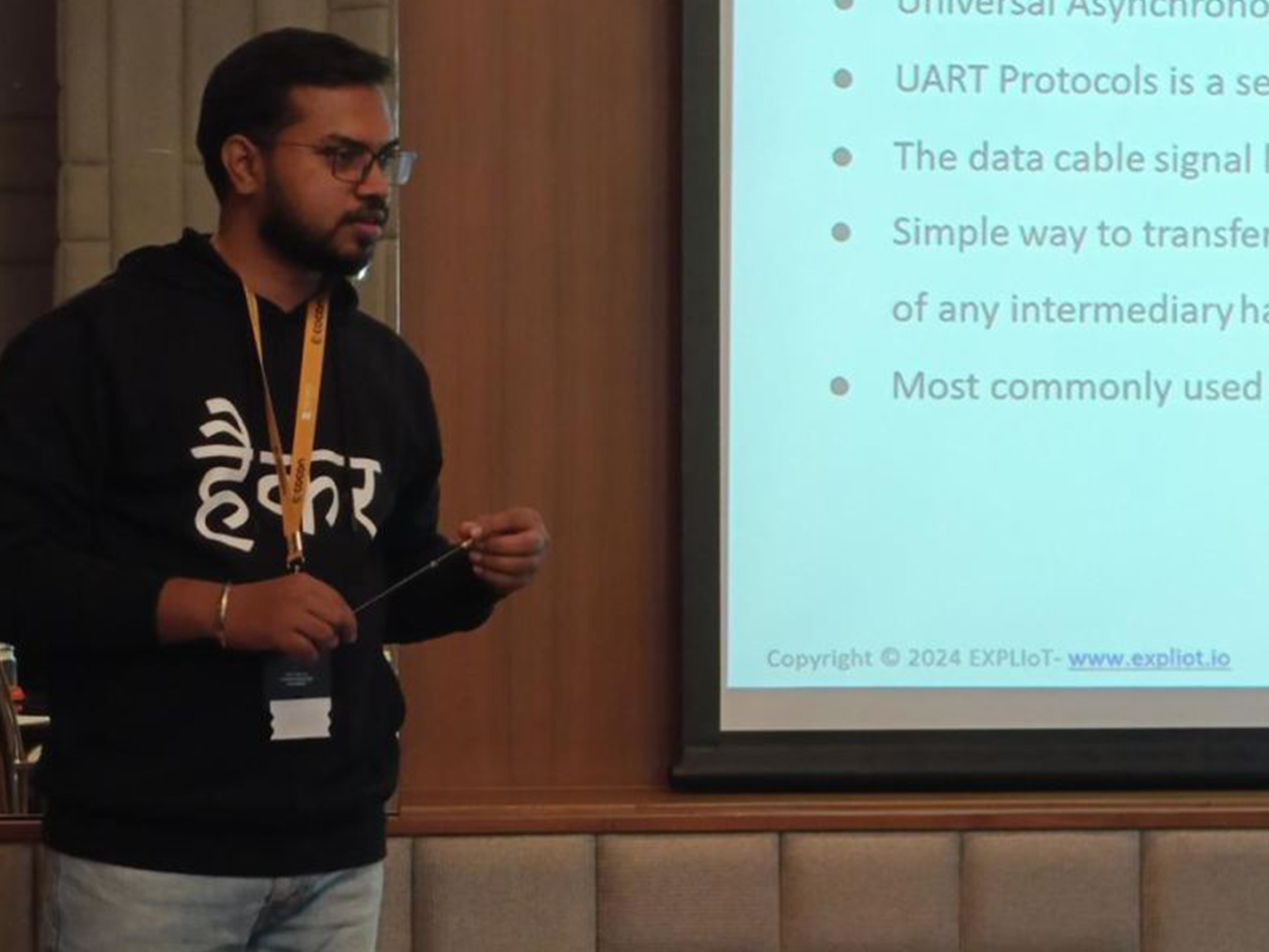A man wearing glasses, a black hoodie with Hindi text, and a yellow lanyard is giving a presentation while holding a pointer stick; a slide projected on the right side partially shows text about UART protocols and a copyright notice for EXPLIoT 2024 with a website link.