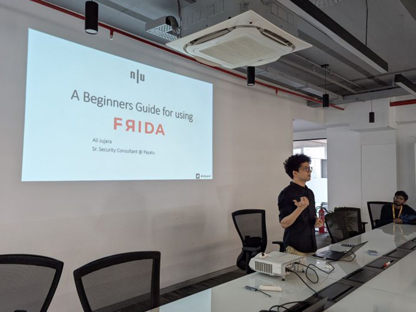 A man stands beside a conference table giving a presentation titled 'A Beginners Guide for using FRIDA' projected on a wall, with a laptop and projector on the table.