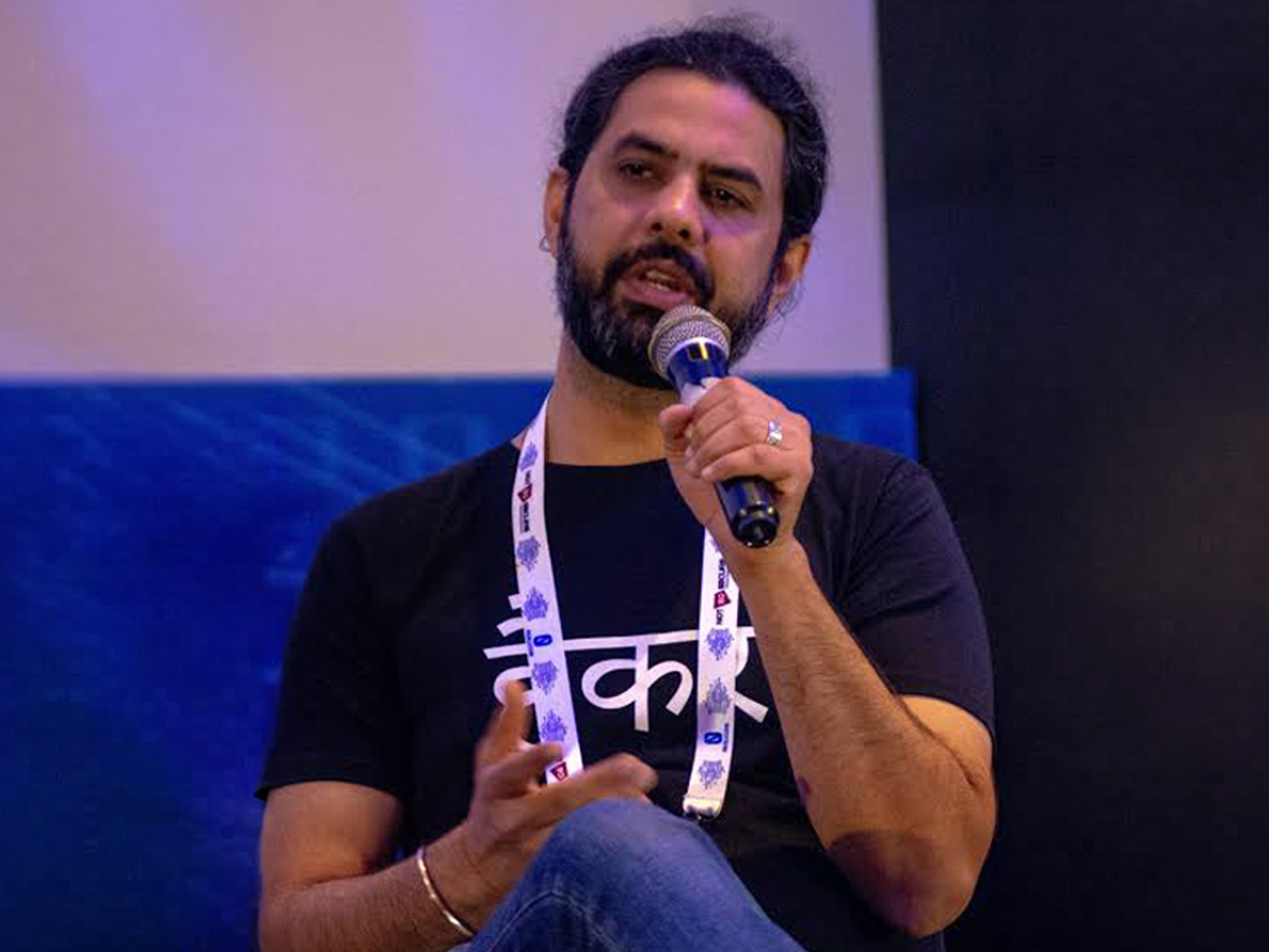 Man with beard and dark hair tied back speaking into a microphone, wearing a black t-shirt with white script and a white lanyard, seated against a blue and black background.