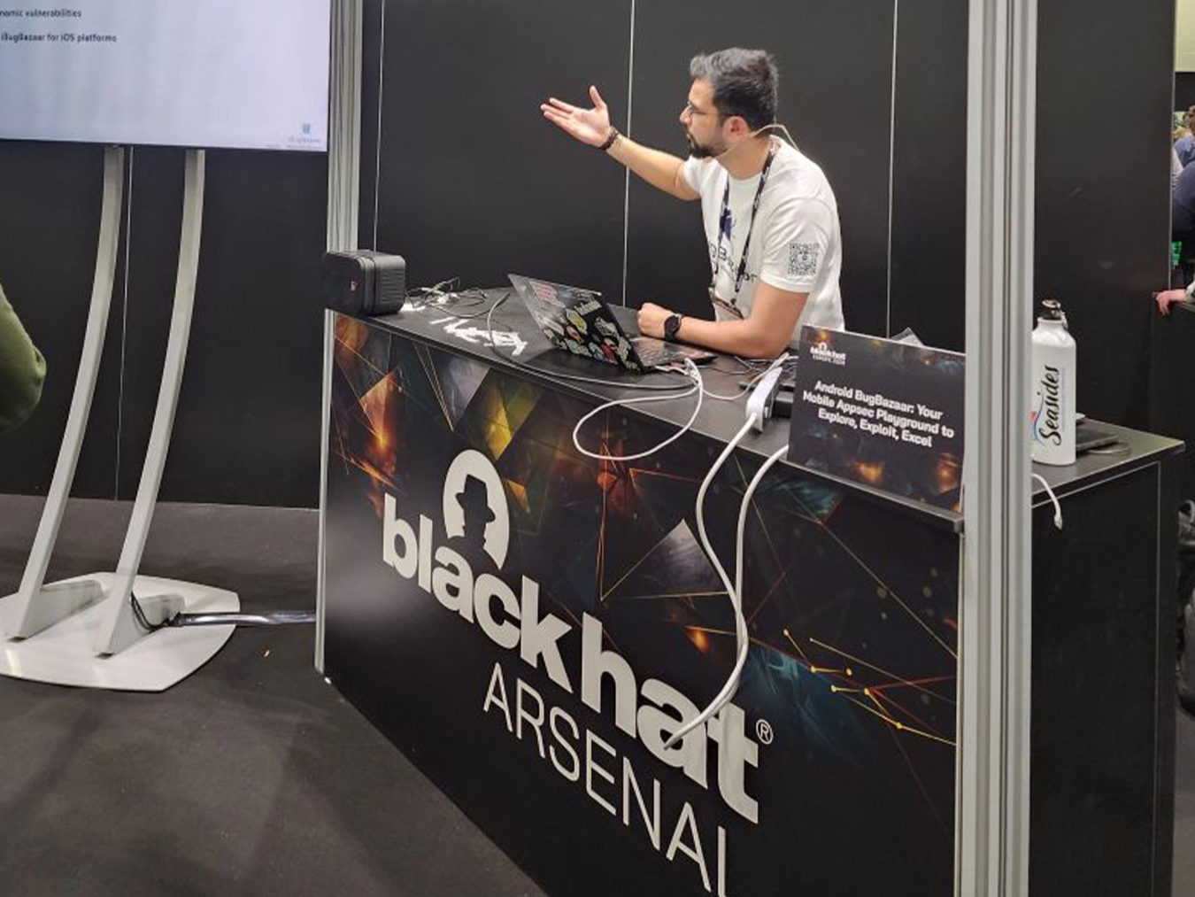 Man with a headset microphone standing behind a Black Hat Arsenal booth, gesturing with his right hand while demonstrating on a laptop covered with stickers, with a sign that reads 'Android BugBazaar: Your Mobile Apps Playground to Explore, Exploit, Excel'.