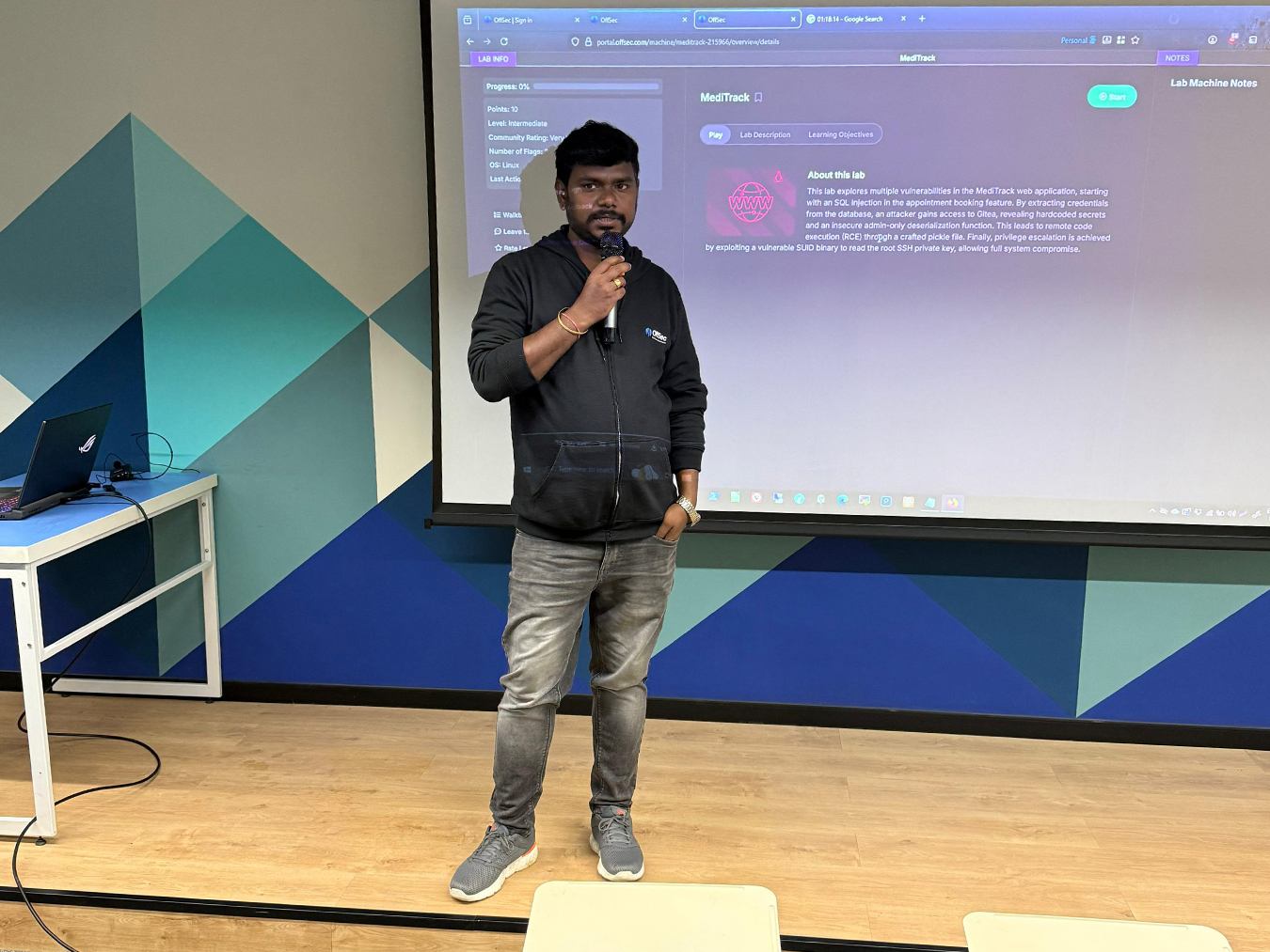 Man with dark hair and beard wearing a black OffSec hoodie and grey jeans, holding a microphone and standing in front of a projector screen displaying a webpage about a Meditrack lab with information on web vulnerabilities and remote code execution.