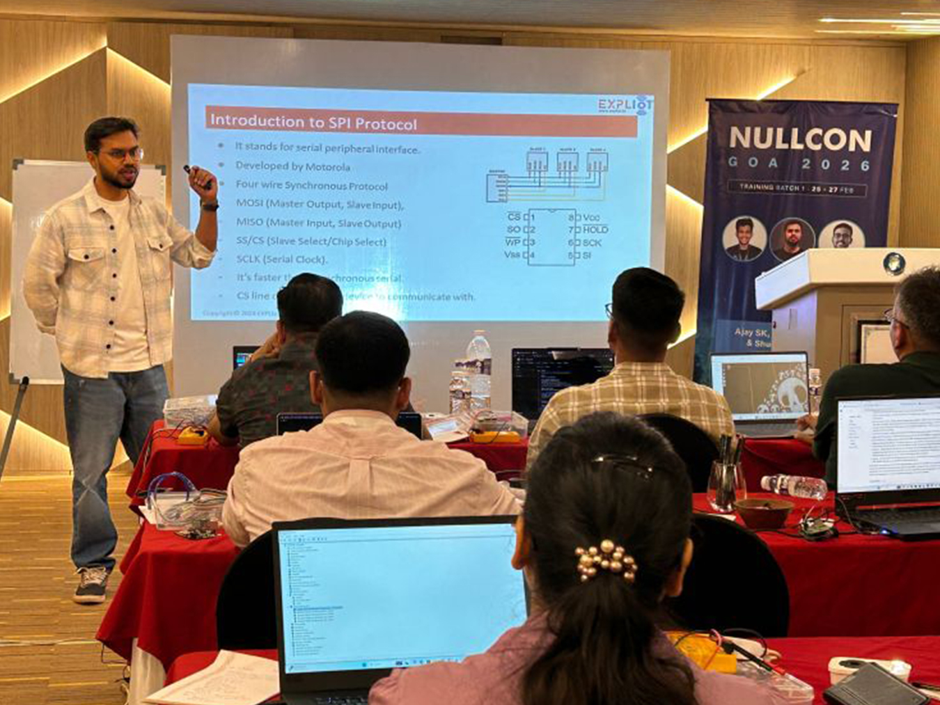 A man giving a technical presentation on the SPI protocol to an audience seated at tables with laptops. The presentation slide explains SPI as a serial peripheral interface protocol developed by Motorola, showing pin diagram and details. A banner on the right side reads 'NULLCON GOA 2026 Training Batch 1: 26-27 Feb' with pictures of three speakers.