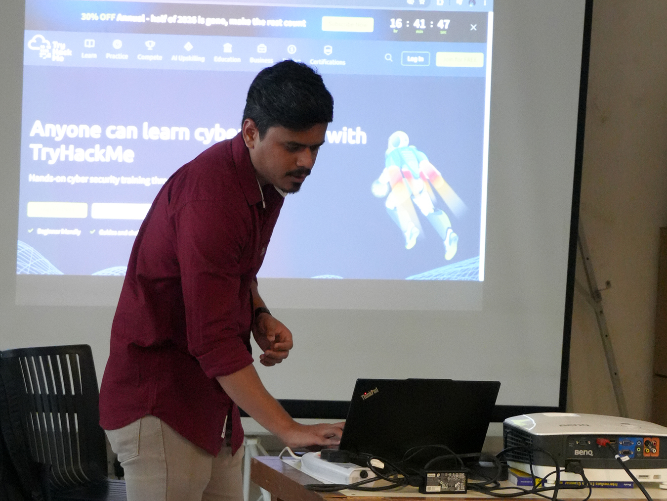 Man in a maroon shirt operating a laptop on a table in front of a projection screen displaying the TryHackMe cybersecurity learning platform homepage.