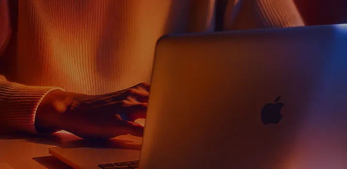 Person wearing an orange sweater typing on a silver Apple laptop in dim lighting.