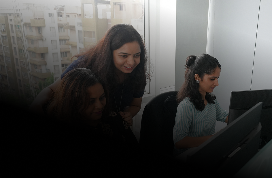 Three women working together in an office, two seated at computer desks while the third stands behind them looking at the screen.