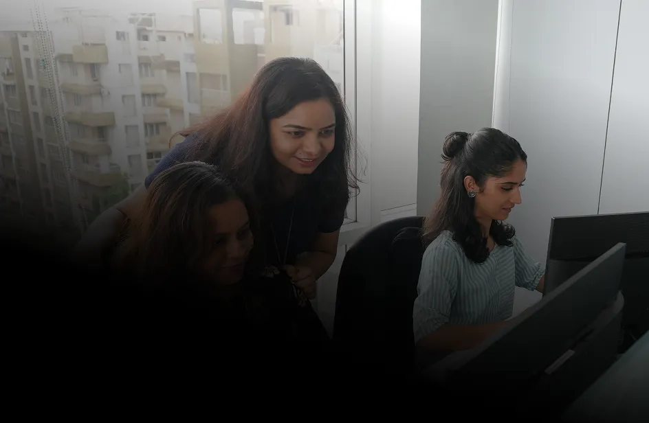 Three women working together in an office, two seated at computer desks while the third stands behind them looking at the screen.