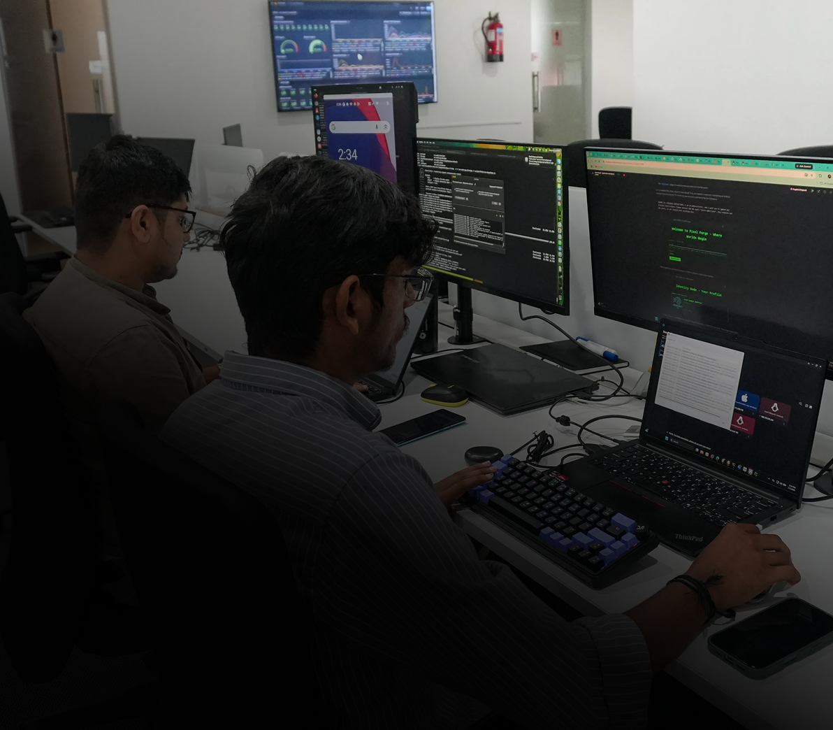 Two men sitting side by side at a desk working on computers in an office, with multiple monitors displaying code and analytics dashboards.