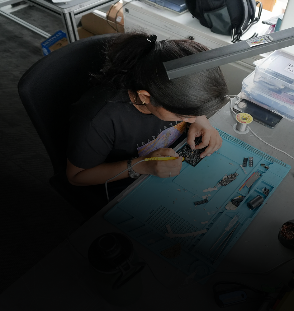 Top view of a person with dark hair tied back, soldering electronic components on a circuit board at a cluttered workstation with tools and materials.