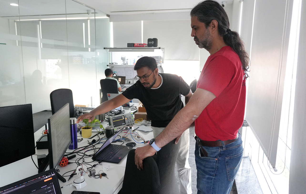 Two men in a modern office workspace focusing on a laptop and electronic equipment on a cluttered white desk with monitors, wires, and gadgets, while another person works on a computer in the background.