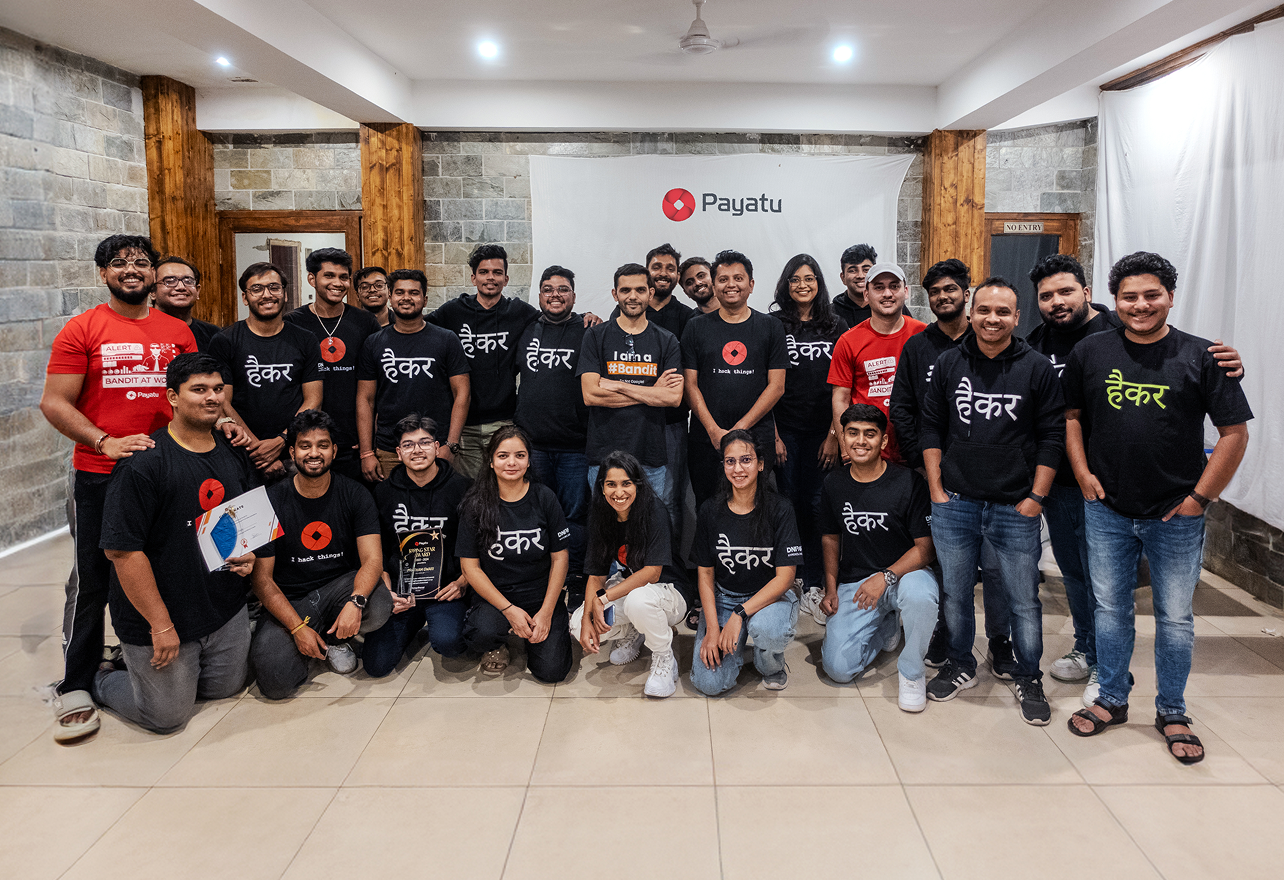 Group photo of 27 people posing indoors in front of a white banner with the Payatu logo, many wearing black t-shirts or hoodies with Hindi text, some holding plaques or certificates, smiling at the camera.