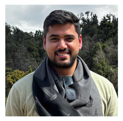 Smiling man with short dark hair and a beard, wearing a beige top and a black scarf with sunglasses hanging from it, standing outdoors with a background of trees and a cloudy sky.