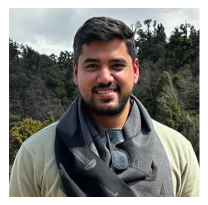 Smiling man with short dark hair and a beard, wearing a beige top and a black scarf with sunglasses hanging from it, standing outdoors with a background of trees and a cloudy sky.