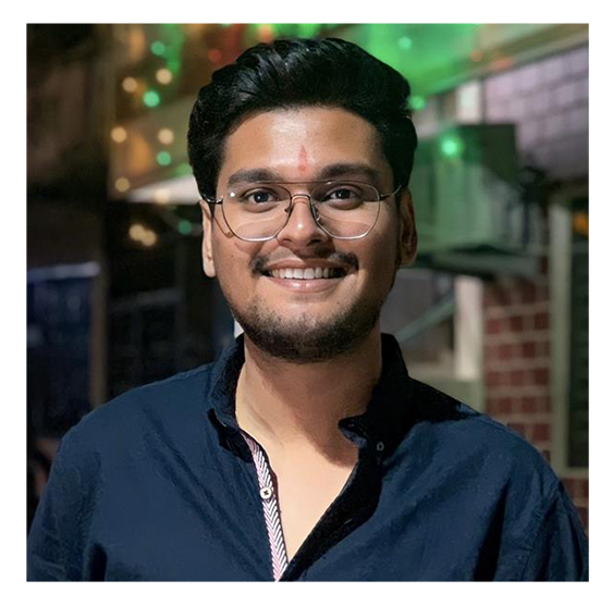 Smiling young man with glasses and dark hair, wearing a navy blue shirt, standing indoors with blurred lights and structures in the background.