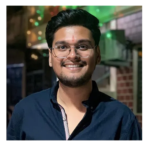 Smiling young man with glasses and dark hair, wearing a navy blue shirt, standing indoors with blurred lights and structures in the background.