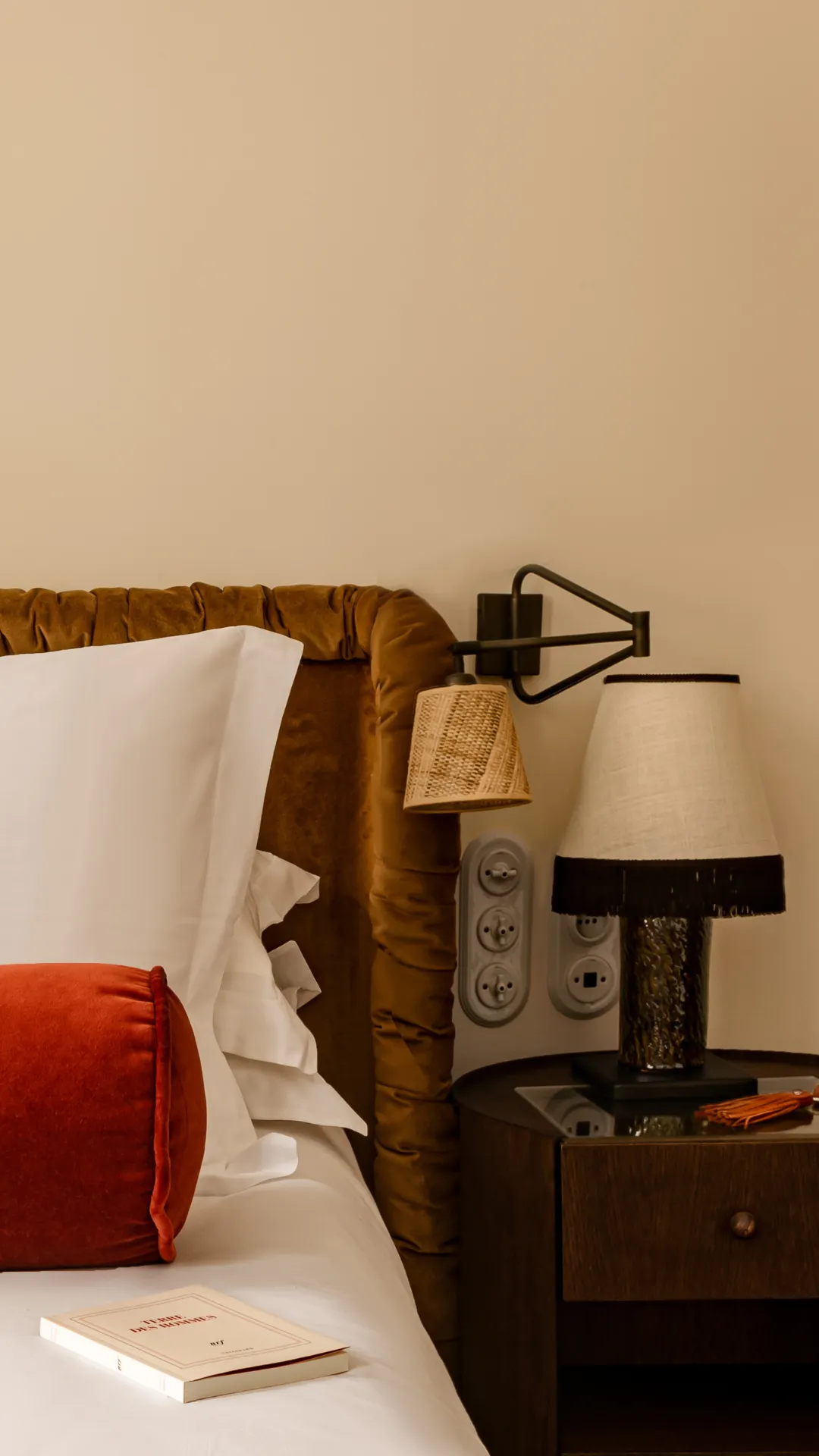 Une tête de lit velours marron, un coussin orange, un livre sur un lit blanc près d'une table de nuit.