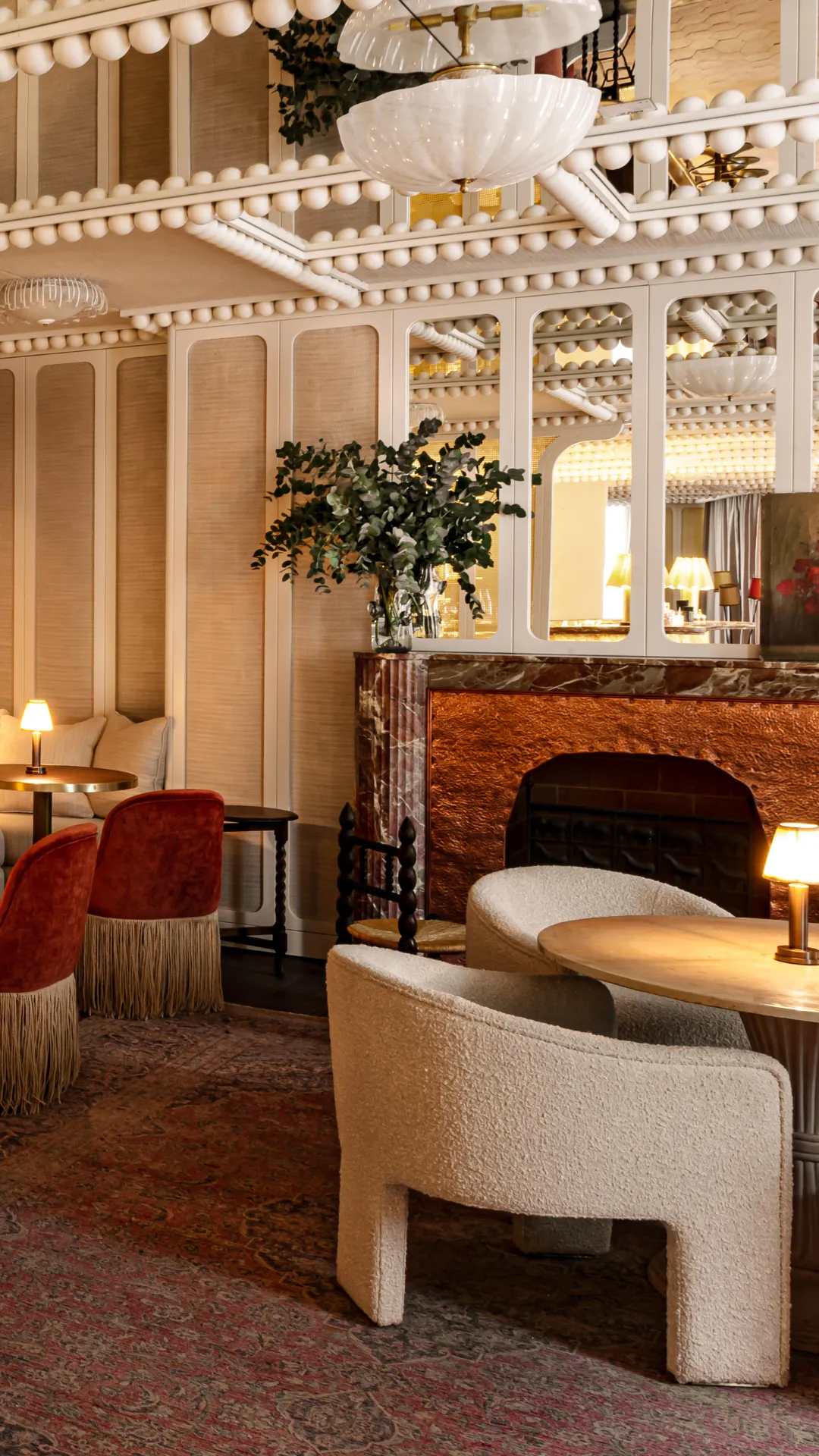 Salon élégant avec fauteuils crème, chaises rouges, cheminée en marbre et grandes miroirs.