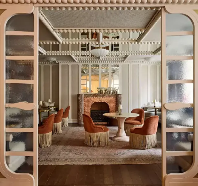 Salle élégante avec des fauteuils orangés à franges autour de tables blanches près d’une cheminée décorative.