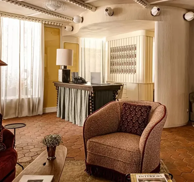 Salon élégant avec fauteuil marron, table basse en bois, bureau rétro et grande fenêtre avec rideaux blancs.