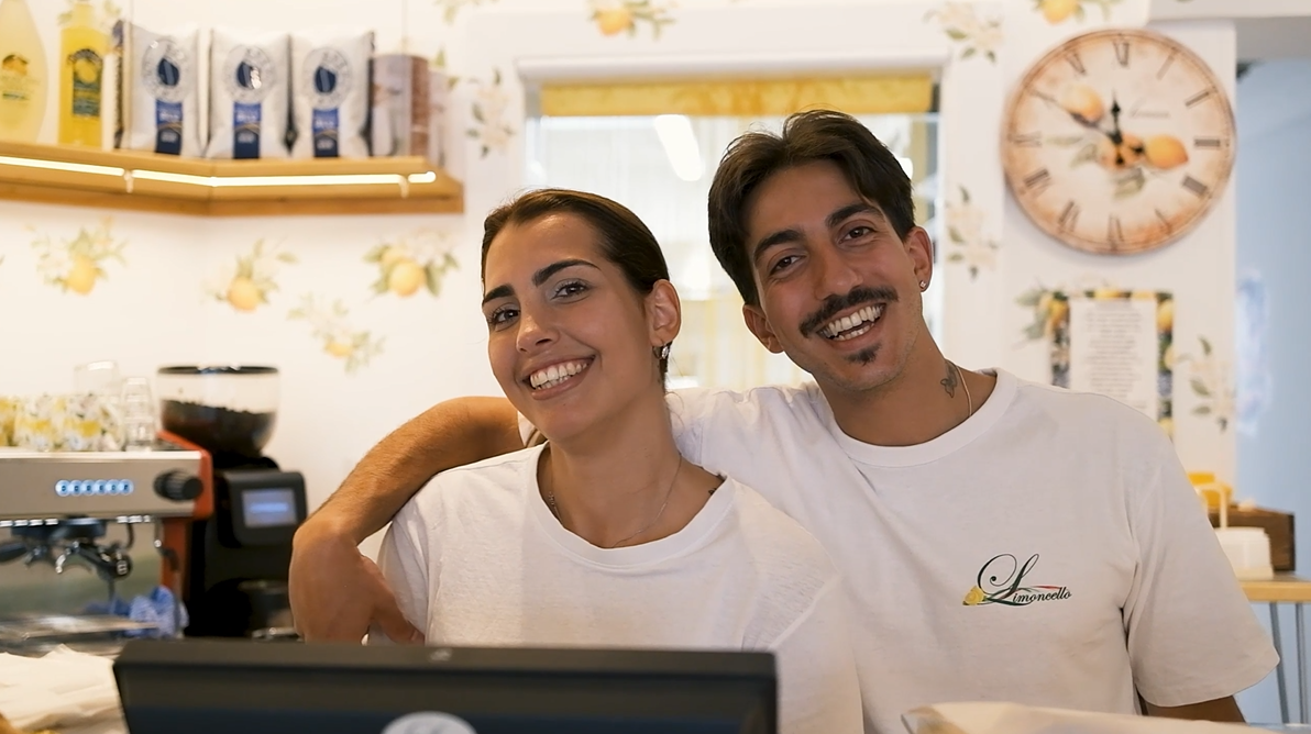 Two smiling young adults, a woman and a man, standing closely together behind a counter in a bright café with lemon-themed decor, including wallpaper and a clock, the man has his arm around the woman.