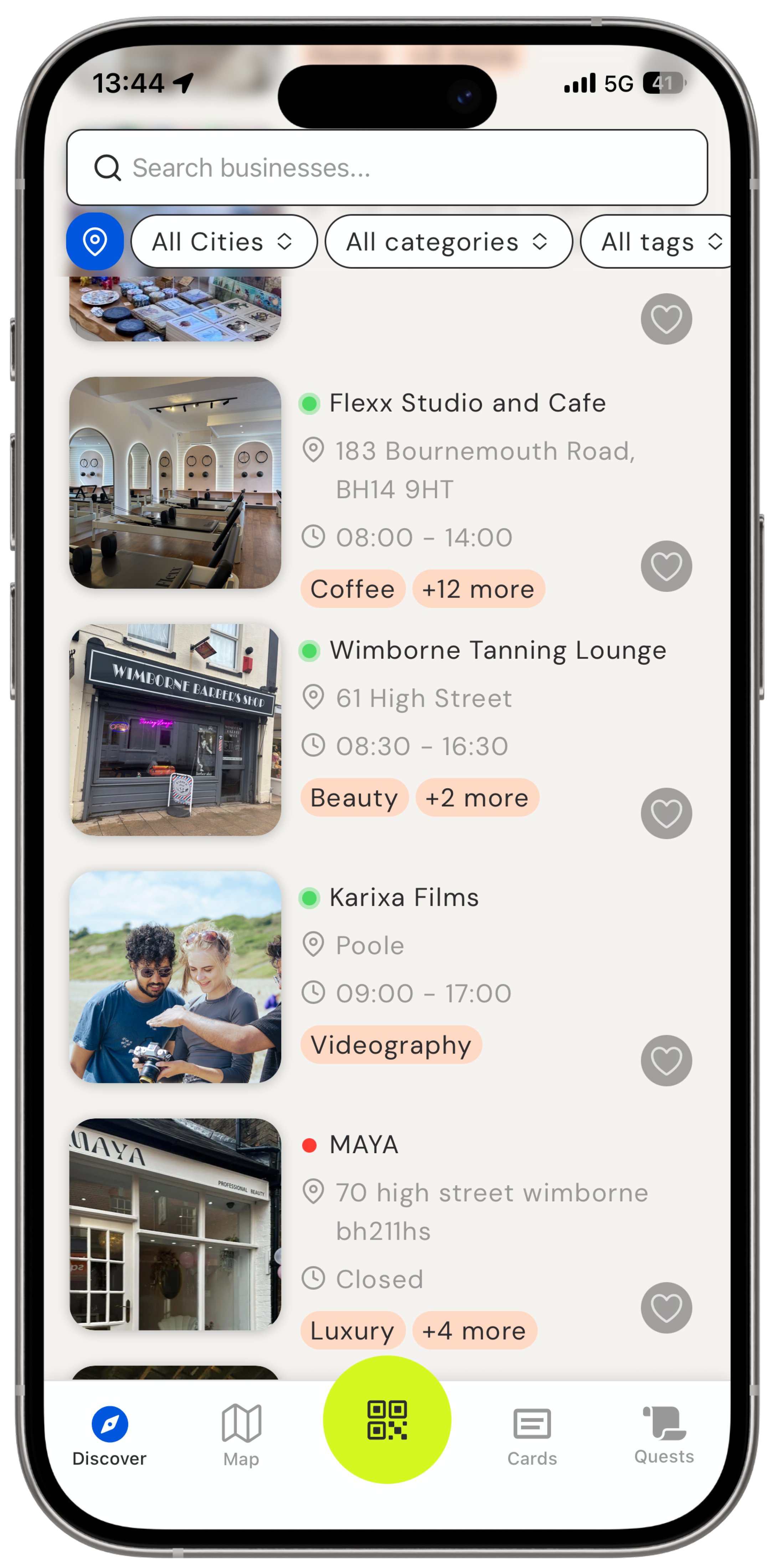Mobile phone screen showing a business directory app with a search bar and filters for cities, categories, and tags. Four business listings are visible: Flexx Studio and Cafe at 183 Bournemouth Road open 08:00-14:00 with coffee and other options; Wimborne Tanning Lounge at 61 High Street open 08:30-16:30 with beauty services; Karixa Films in Poole open 09:00-17:00 offering videography; and MAYA at 70 High Street Wimborne, currently closed, with luxury services. Each listing includes a thumbnail, address, hours, and category tags. The bottom navigation bar has icons for Discover, Map, QR code scanner, Cards, and Quests.