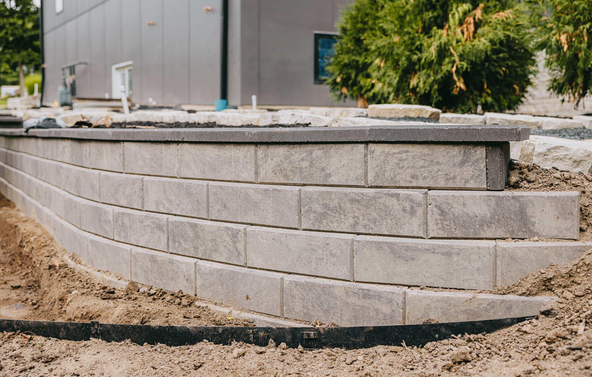 Curved concrete block retaining wall under construction on freshly graded clay beside a modern home.