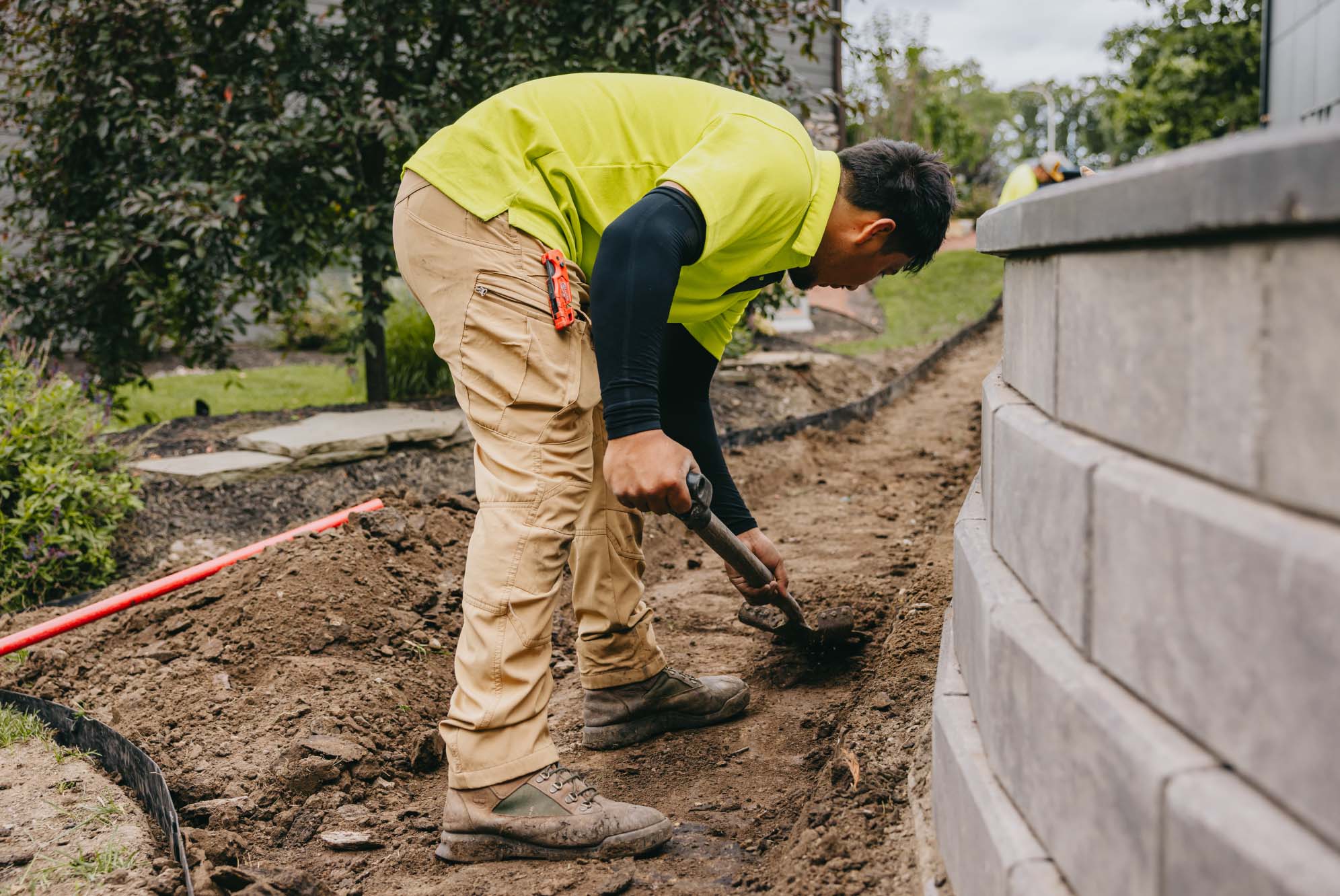 Crew member trenching by hand beside a new stone wall, with utility locate paint marking the ground.