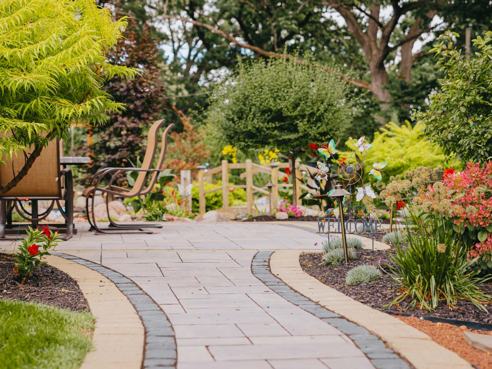 Brick paver walkway curving past a seating area framed by deep green planting.