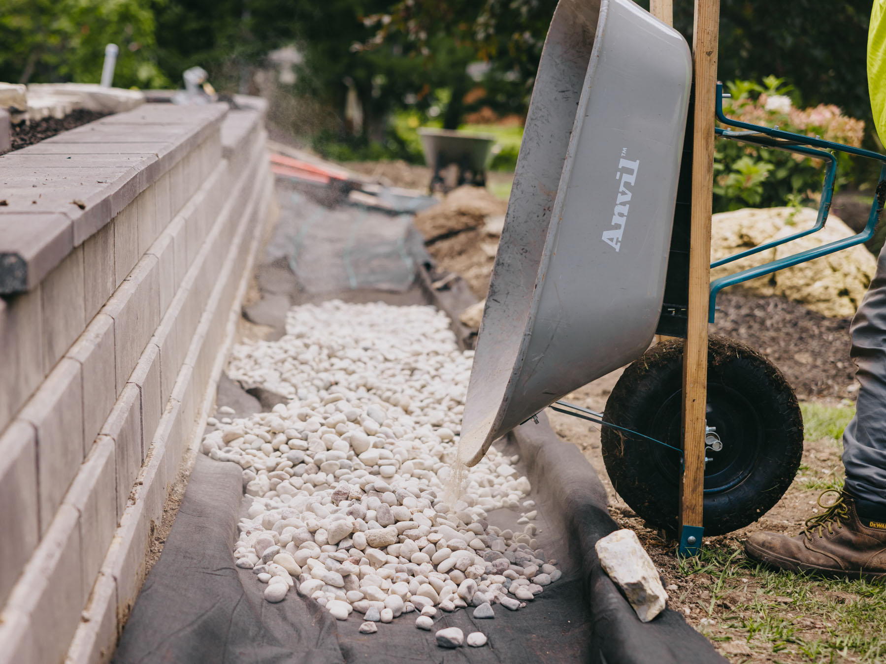 Crew member pouring decorative river rock along the base of a new retaining wall.