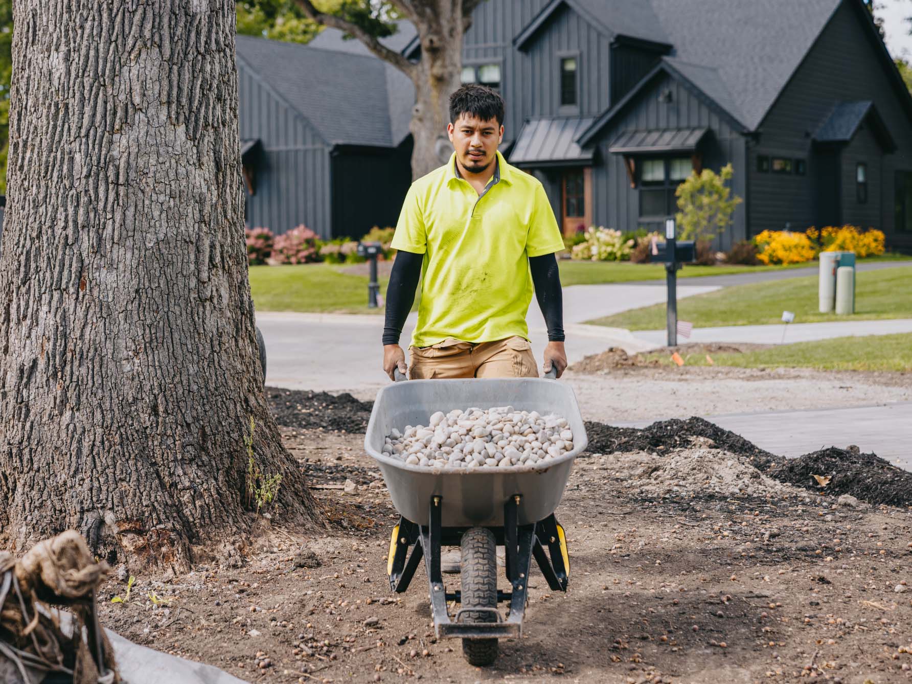 Crew member moving a wheelbarrow of river rock through a garden under construction.