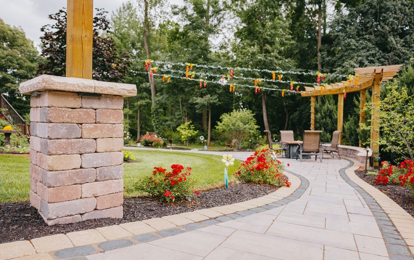 Wood pergola over a paver patio with flower beds in a suburban backyard.
