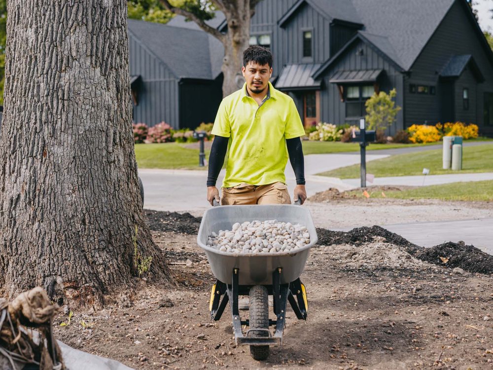 Crew member moving a wheelbarrow of river rock through a garden under construction.