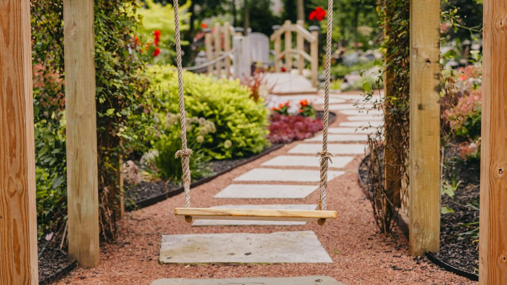 Wooden garden swing looking out over a stone pathway through a deep planted border.